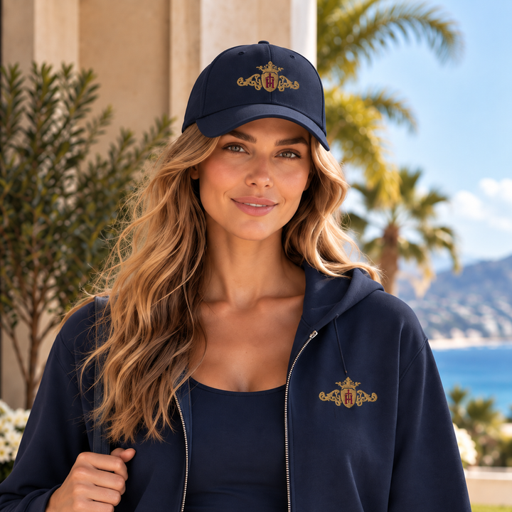 A woman wearing a navy blue baseball cap and matching zip-up hoodie with a bronze-gold and maroon crest embroidery, posed outdoors with a beach and palm tree background.