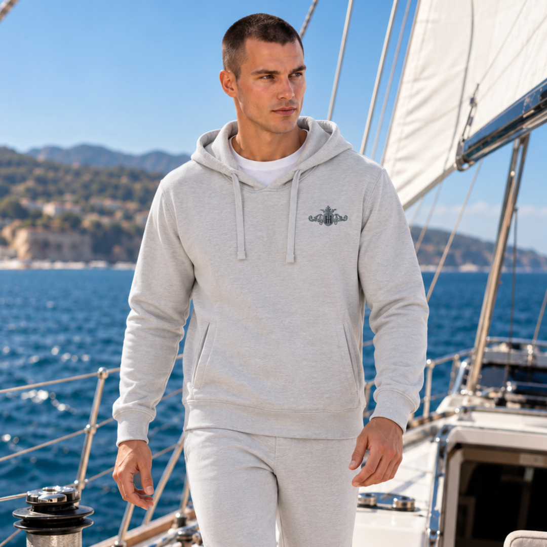 A man wearing a light gray heathered side pocket hoodie and matching sweatpants stands on a sailboat with the blue sea and coastline in the background. The hoodie features a small dark graphic on the left chest.