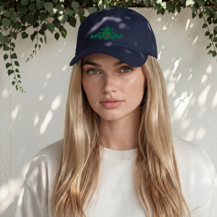 Woman wearing a navy blue cap with a an embroidered kelly-green logo against a white wall with greenery.