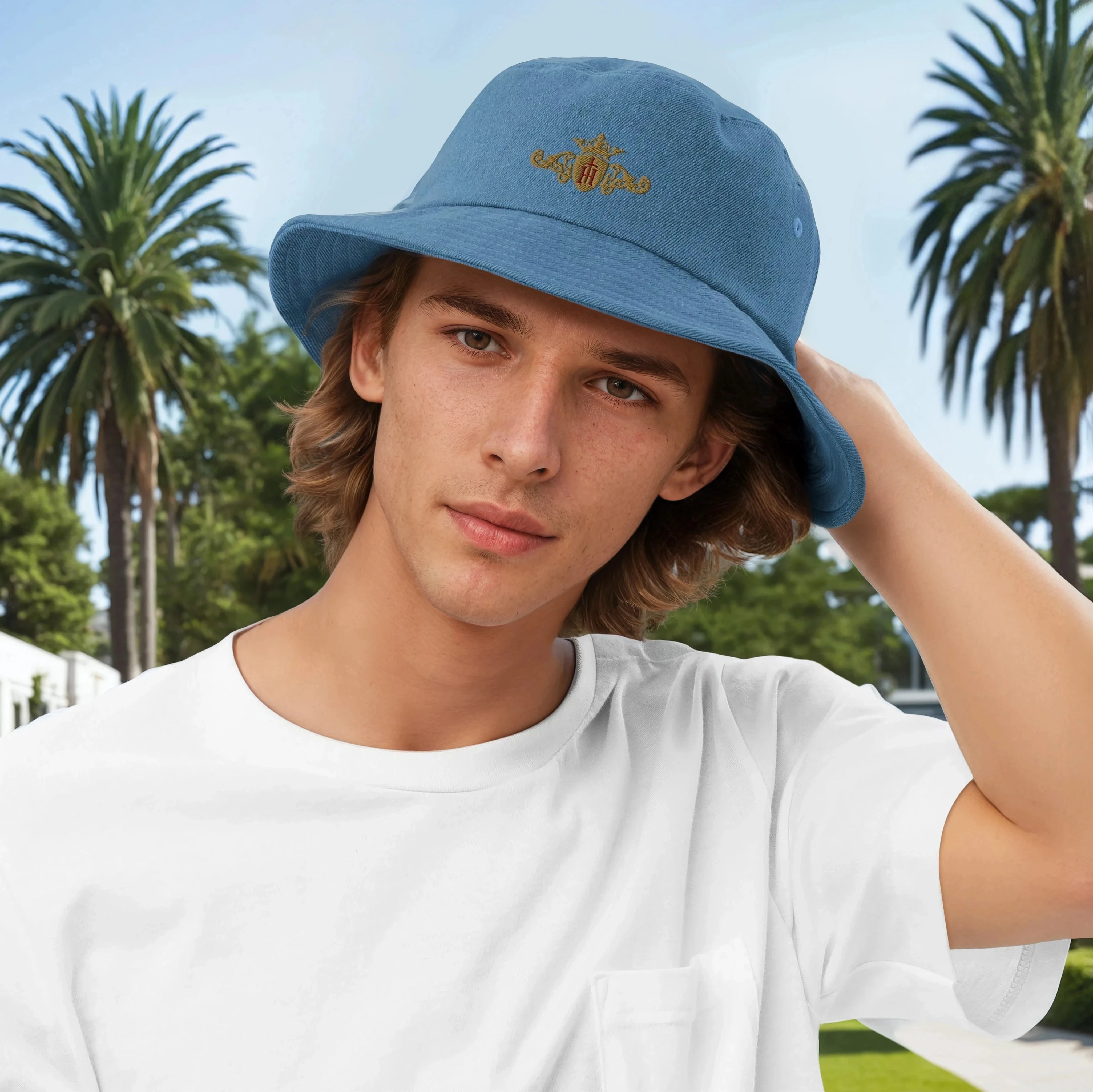 Young man wearing a blue bucket hat with Helen Of Troy Apparel Beverly Hills  gold crest with a maroon center, standing outdoors with palm trees in the background.