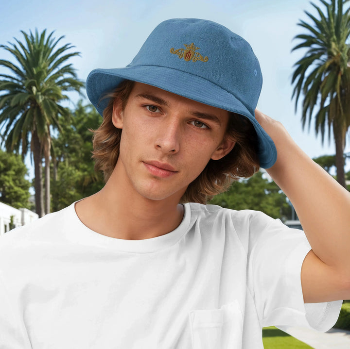 Young man wearing a blue bucket hat with Helen Of Troy Apparel Beverly Hills  gold crest with a maroon center, standing outdoors with palm trees in the background.