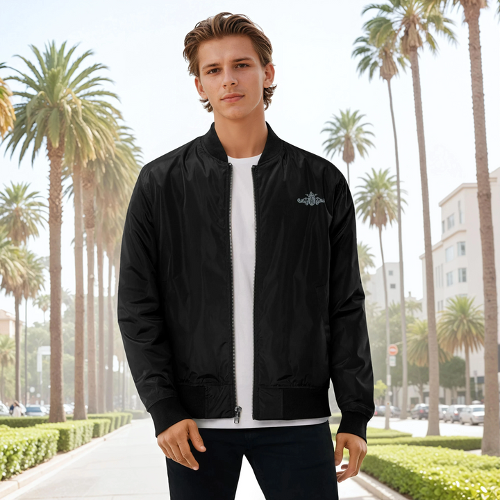 Man wearing a black bomber jacket with a grey and black crest in front of a palm tree-lined street in the background