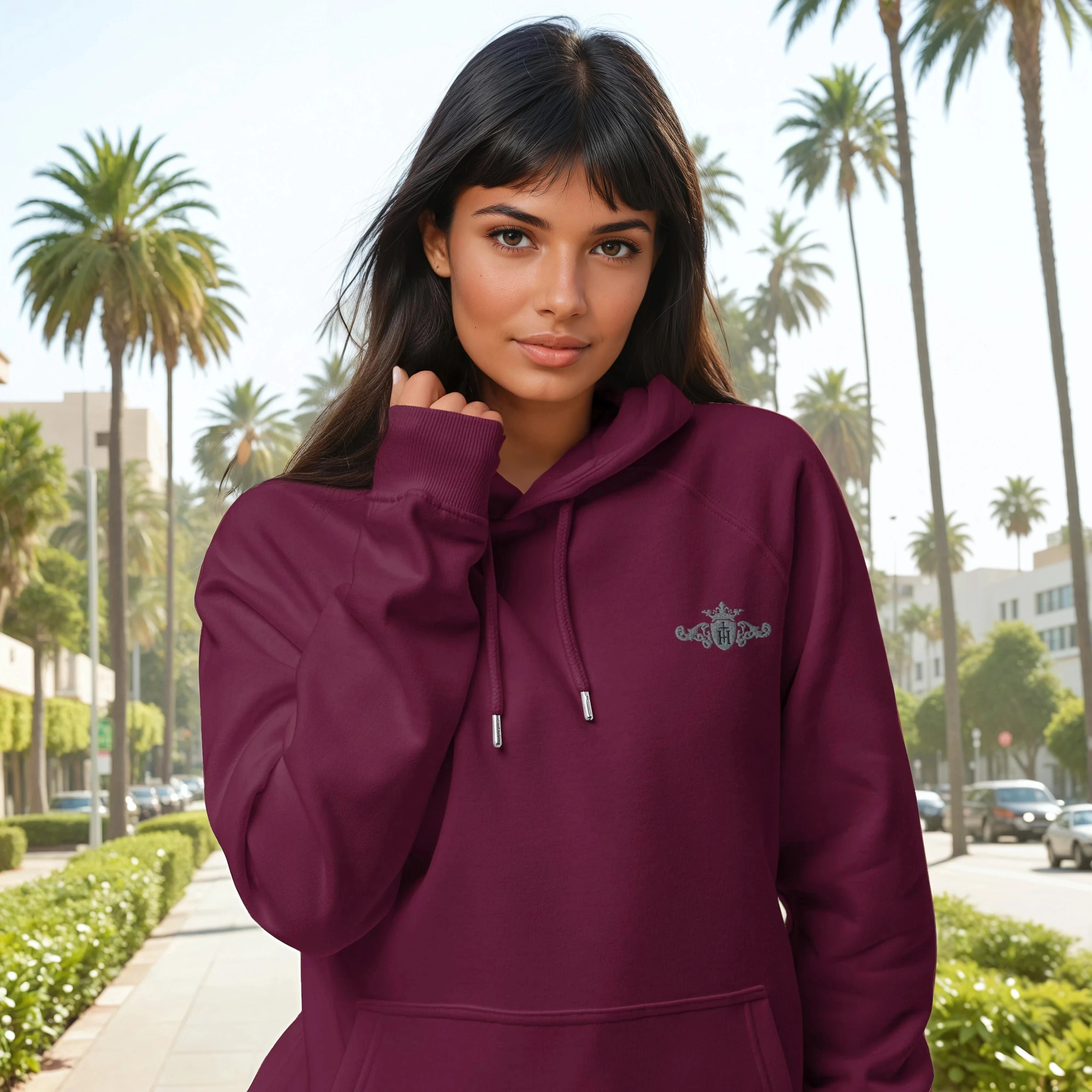 Woman wearing a maroon hoodie with a grey Helen Of Troy Apparel Beverly Hills crest with a black center in front of a palm tree-lined street.